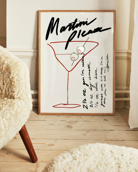 The Martini Please Poster, displayed on a wooden floor, features a stylish illustration of a martini glass with olives alongside handwritten measurements and instructions for a classic martini recipe. A fuzzy chair in the foreground enhances the cozy ambiance.