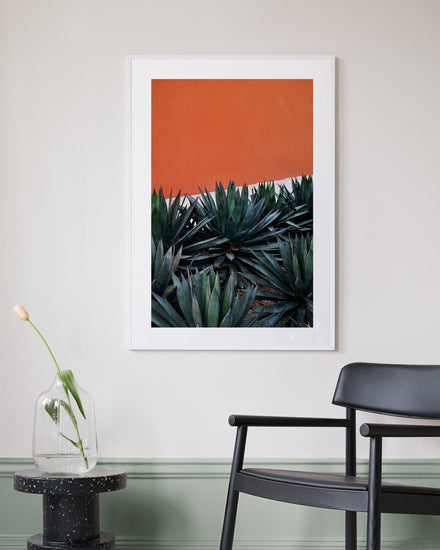 The Pershing Square Red Poster, featuring green agave against an orange wall, evokes a Los Angeles vibe as it hangs on a light gray wall. Nearby, a black chair is next to a round black table with a clear vase and pink tulip. The wall base is painted green.