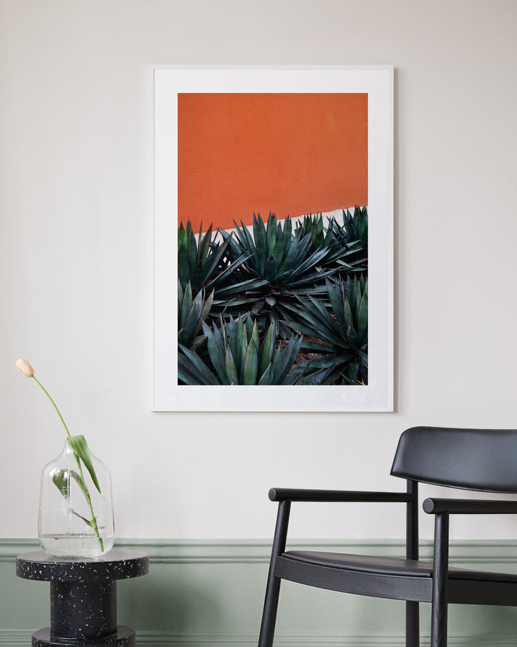 The Pershing Square Red Poster, featuring green agave against an orange wall, evokes a Los Angeles vibe as it hangs on a light gray wall. Nearby, a black chair is next to a round black table with a clear vase and pink tulip. The wall base is painted green.