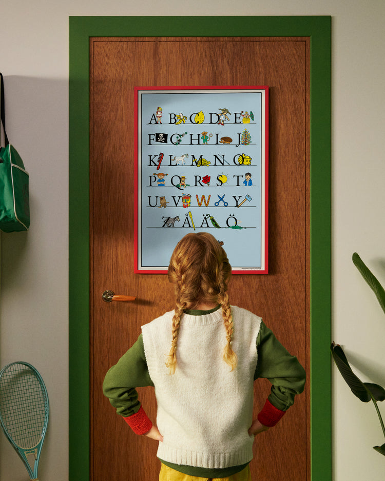 A child standing in front of a door looking at a Pippi Longstocking Swedish alphabet poster A–Ö.