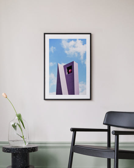 A modern, minimalist room features a framed Pershing Square Tower Poster depicting a tall, purple structure against a blue sky. A black chair is next to a small black table, supporting a glass vase with one drooping tulip.