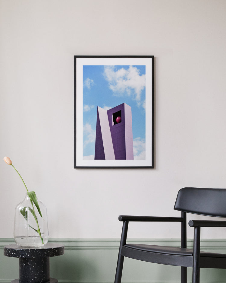 A modern, minimalist room features a framed Pershing Square Tower Poster depicting a tall, purple structure against a blue sky. A black chair is next to a small black table, supporting a glass vase with one drooping tulip.