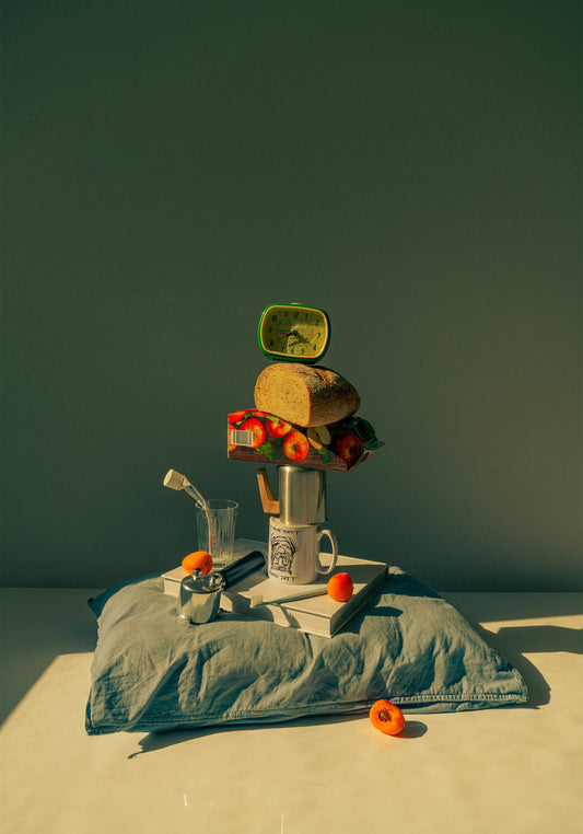 A still life featuring various breakfast elements including a slice of bread, apple juice, coffee mug, teapot, with a pillow underneath and an alarm clock on top.