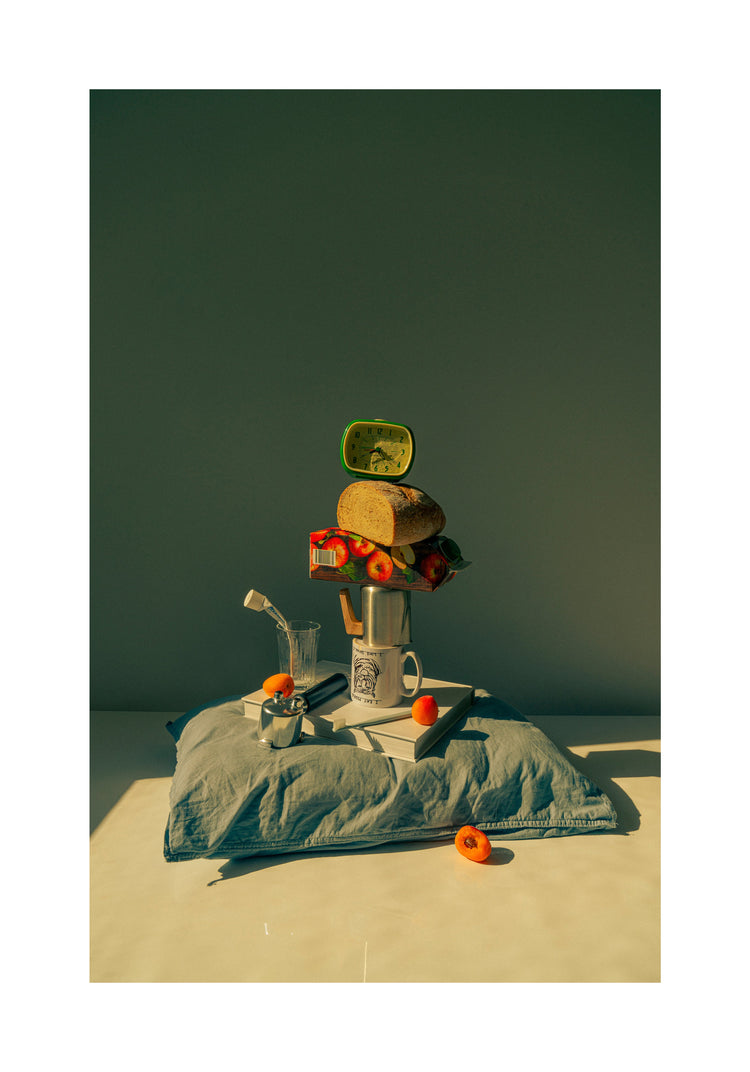 A still life featuring various breakfast elements including a slice of bread, apple juice, coffee mug, teapot, with a pillow underneath and an alarm clock on top.