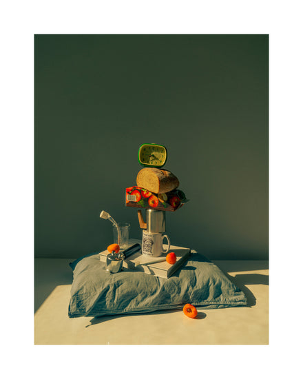 A still life featuring various breakfast elements including a slice of bread, apple juice, coffee mug, teapot, with a pillow underneath and an alarm clock on top.