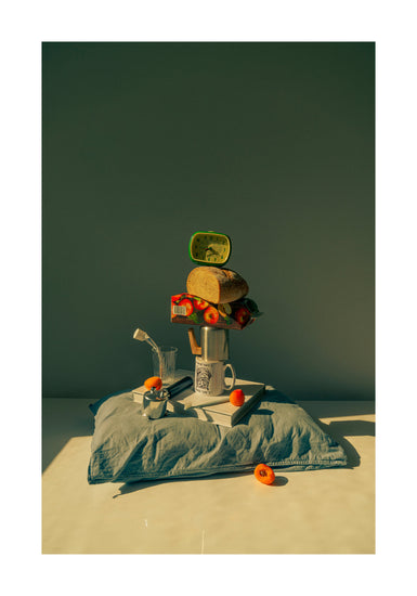 A still life featuring various breakfast elements including a slice of bread, apple juice, coffee mug, teapot, with a pillow underneath and an alarm clock on top.