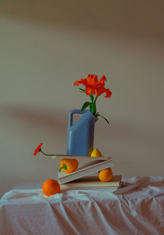 The Hanging in There Poster showcases a still life with a blue jug, red flowers, stacked books, and scattered oranges and pears on a white cloth. Vibrant colors create an artistic ambiance that evokes the warmth of Berlin in winter against a plain backdrop.