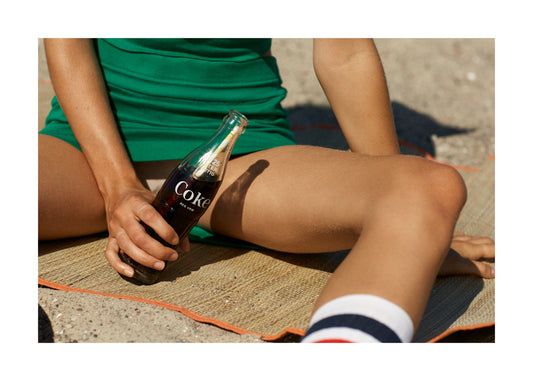 On a sunny beach, someone in a green outfit relaxes on a mat with a glass Coke bottle, their shadowy legs capturing the nostalgic charm of retro girl posters. The sandy background adds to its appeal as wall art. Inspired by the Girls Just Want To Chill Out Poster.