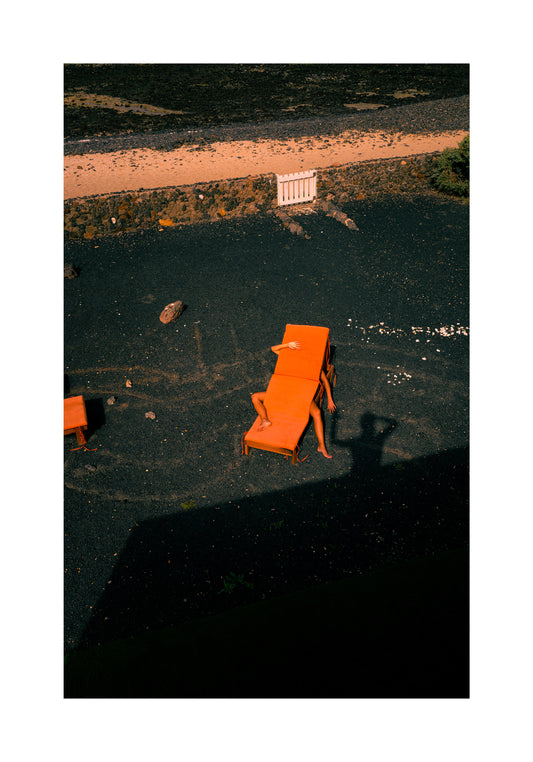 A person relaxes on an orange sunbed amid dark gravel, under partial building shade. Another chair awaits nearby. Long shadows, like a silent wave, stretch across the ground in The Short Romantic Story Between a Chair and a Shadow poster.