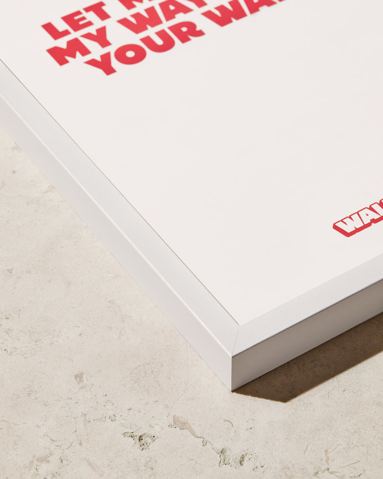 Close-up of a poster in the Frame White on a light-textured surface. The poster displays large red text, partially visible, with the phrase starting with LET and ending with YOUR WAY.