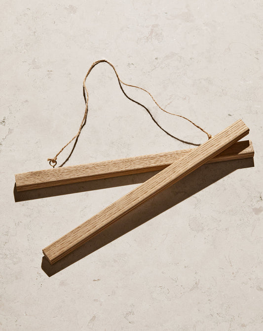 The Poster hanger Oak consists of two solid oak wooden bars with a thin rope connection and discreet magnets to secure your artwork. They lie parallel on a textured, light surface, showcasing a natural wood grain.