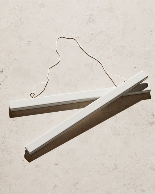 Two Poster hanger White pieces crafted from solid pine, complete with an attached string, rest on a light, textured surface. Sunlight casts soft shadows that highlight their minimalist design and the discreet magnets that effortlessly secure your art.