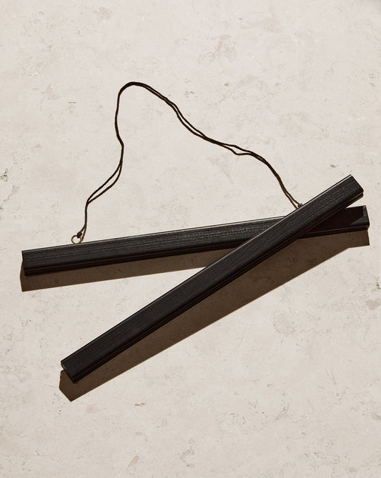 A Poster hanger Black with two sleek black wooden sticks and a string attached lays diagonally on a light marble surface, showcasing its thin rectangular shape and looped string design.