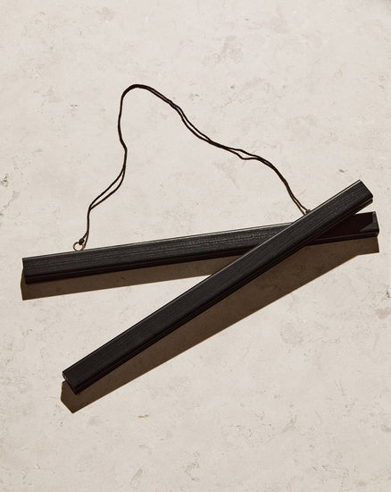 A Poster hanger Black with two sleek black wooden sticks and a string attached lays diagonally on a light marble surface, showcasing its thin rectangular shape and looped string design.