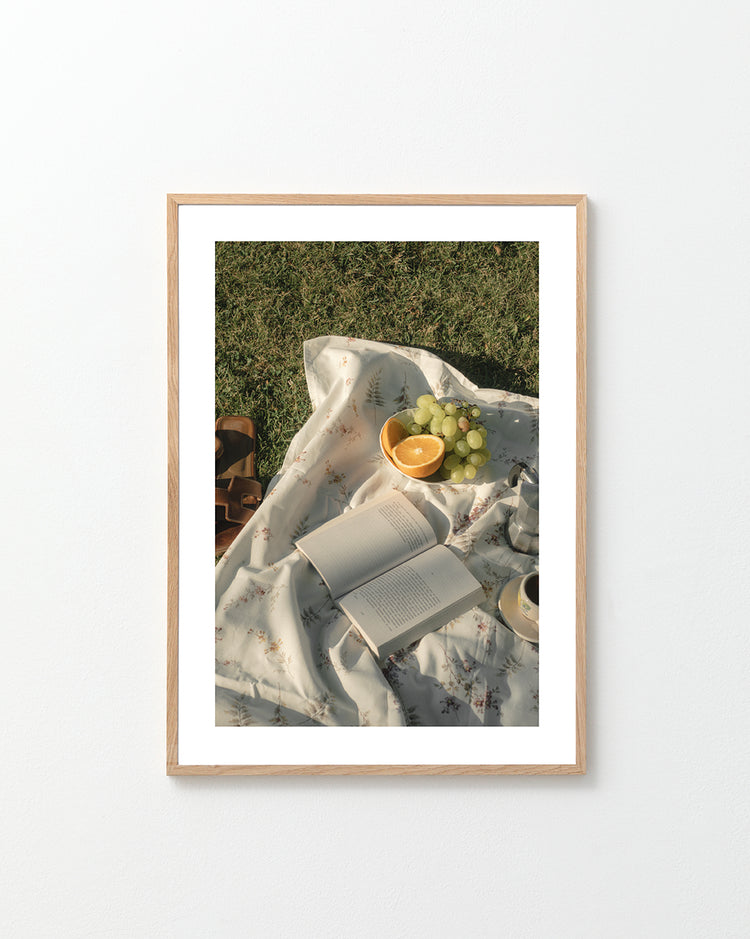 The Tuscan Summer Poster captures a serene picnic on lush grass with a floral blanket cradling an open book, sliced oranges, grapes, and a cup. Brown sandals add to the relaxed outdoor vibe, perfect for inviting moments under the sky.