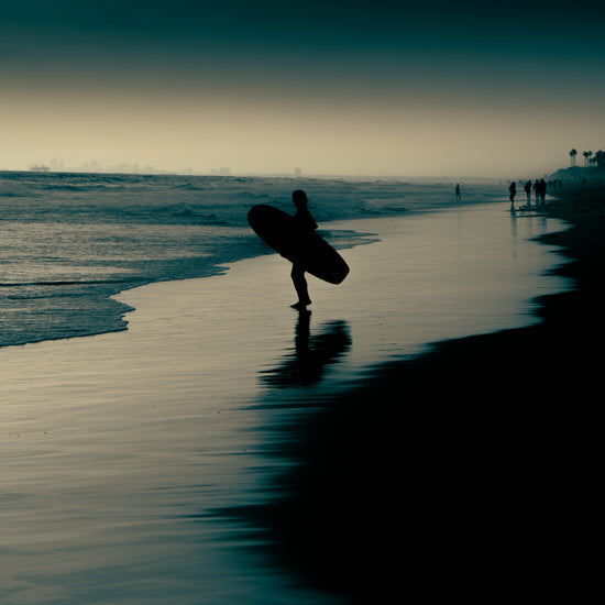 Surfer standing on the shore during low tide under a darkening sky.