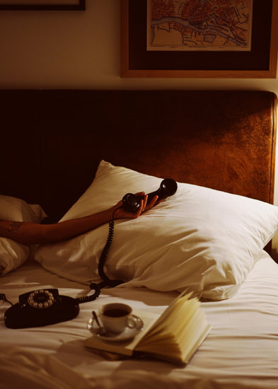 In a dimly lit bedroom, a hand holds an old-school telephone receiver resting on a pillow, surrounded by an open book, coffee cup, and phone base on the bed. Above, the art wall features the Do Not Disturb poster.