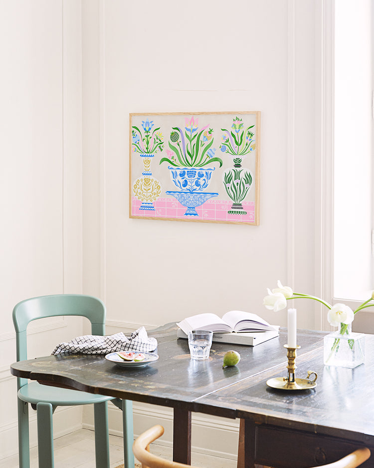 A dining table set against a light wall features the Ceramics Poster, with colorful framed botanical artwork reminiscent of Swedish kurbits. The table displays a green chair, checkered cloth, an open book, a glass of water, vibrant green fruit, and two white flowers in a brass candlestick holder.