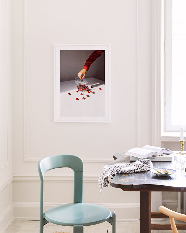 A minimalistic room features a teal chair and dark wooden table. The Summer Treats poster on the wall displays a hand with a red sleeve holding a bouquet above white, scattered with red petals. Natural light streams through a window on the right.