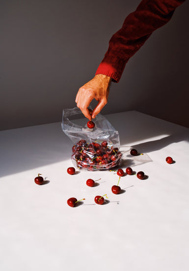 A hand reaches into an open bag of cherries on a white surface in the Summer Treats Poster, with scattered cherries and lighting that highlights their deep red color against strong shadows.