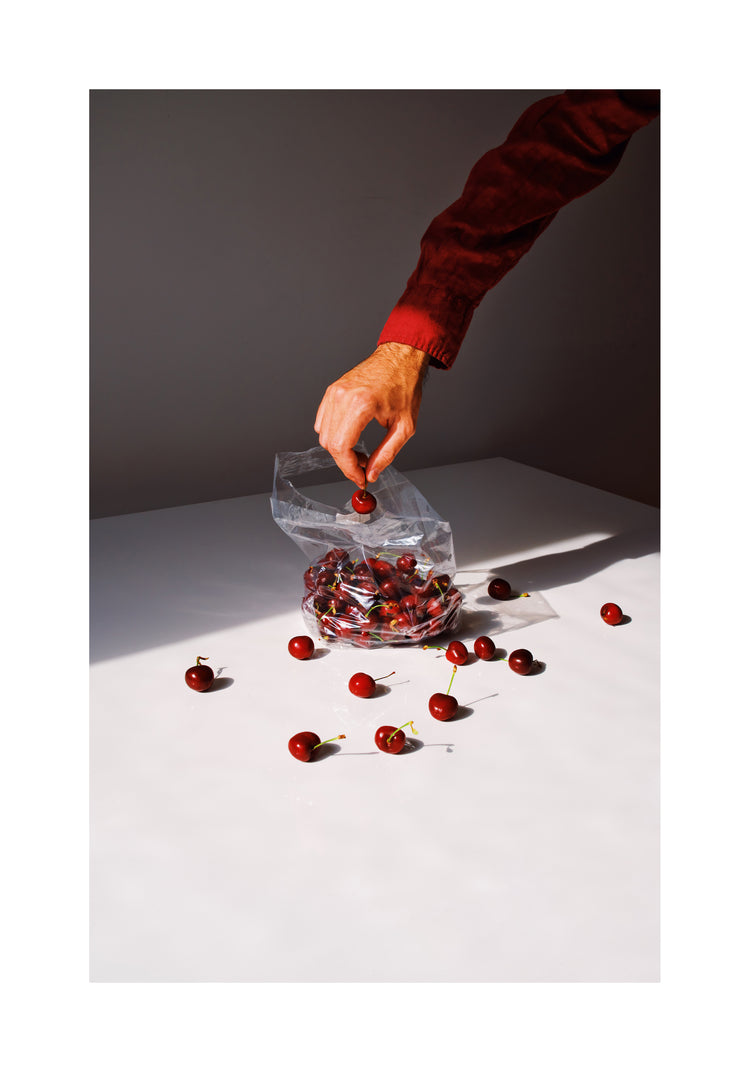 A hand in a red long-sleeved shirt reaches for cherries inside a clear bag on a white surface. Loose cherries scatter around, with light and shadows adding drama. Introducing the Summer Treats Poster.