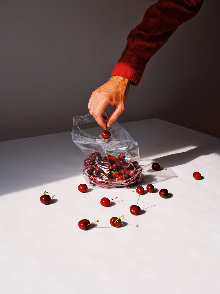 A person in a long-sleeve red shirt reaches into a transparent plastic bag filled with cherries, some scattered on the white table, as depicted in the Summer Treats Poster.