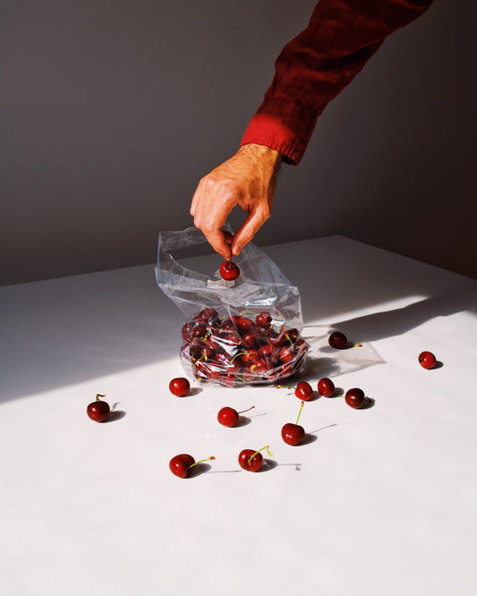 In bright lighting, a hand in a red sleeve reaches for a cherry from an open clear plastic bag filled with cherries, placed on a white surface. Several cherries are scattered around, casting distinct shadows. Summer Treats Poster.