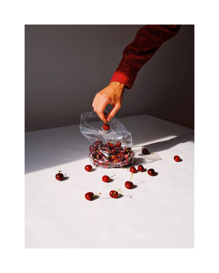 A hand in a red sleeve reaches into a clear cherry bag on a white surface, with scattered cherries casting soft shadows. Bright lighting accentuates reds and whites. Product: Summer Treats Poster.
