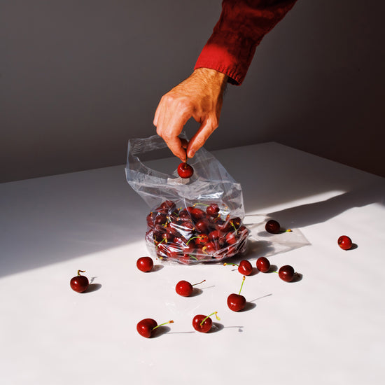 A hand in a red sleeve reaches for a cherry from an open bag on a white surface, with cherries scattered around. Sunlight casts sharp shadows across the scene in this Summer Treats Poster.