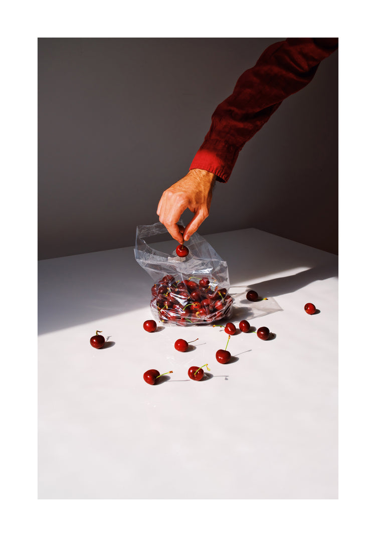 A person in a red long-sleeve shirt reaches into a plastic bag labeled Summer Treats Poster to pick a cherry. Several cherries are scattered on a white surface with strong lighting creating shadows.