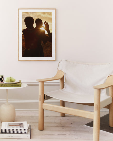 A minimalist living room features a wooden chair and a round side table with books and green apples. The Lets Play Like When We Were Young poster, depicting two silhouetted people waving at sunset, hangs on the light-toned wall.