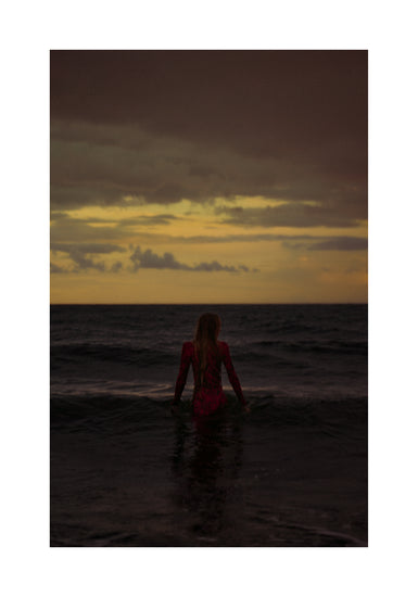 A woman in a red dressing gown walking into the ocean during sunset.