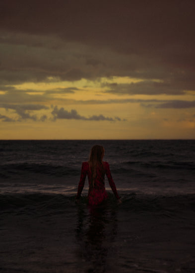 A woman in a red dressing gown walking into the ocean during sunset.