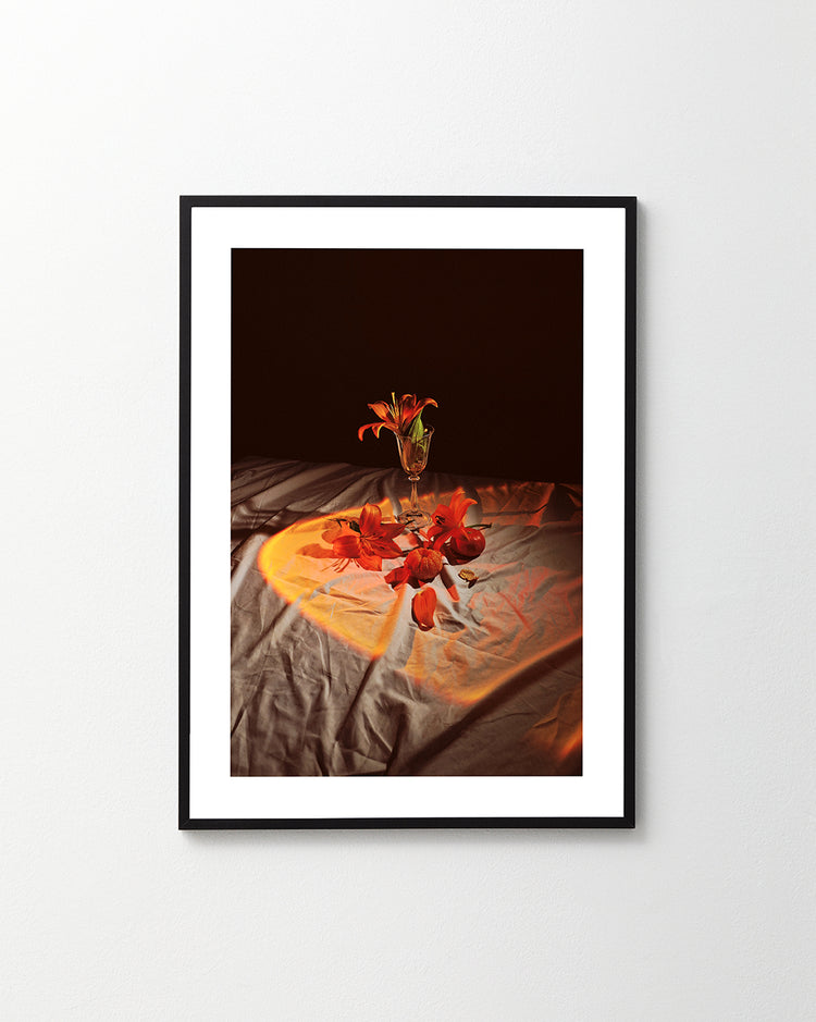 The Insomnia of the Flowers Poster features a framed photograph of a vase with orange-red lilies on draped fabric, softly illuminated and surrounded by scattered petals. Dark background highlights the vibrant flowers, while subtle shadows enhance the composition.