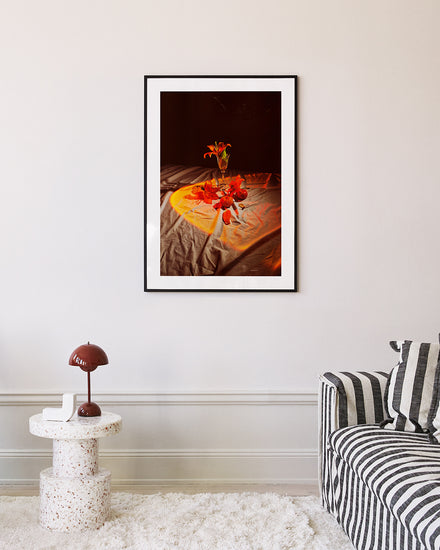 A living room featuring the Insomnia of the Flowers poster above a striped couch. Nearby, a round speckled side table holds a brown mushroom-shaped lamp against a light-colored wall.