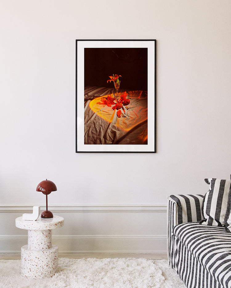 A living room featuring the Insomnia of the Flowers poster above a striped couch. Nearby, a round speckled side table holds a brown mushroom-shaped lamp against a light-colored wall.
