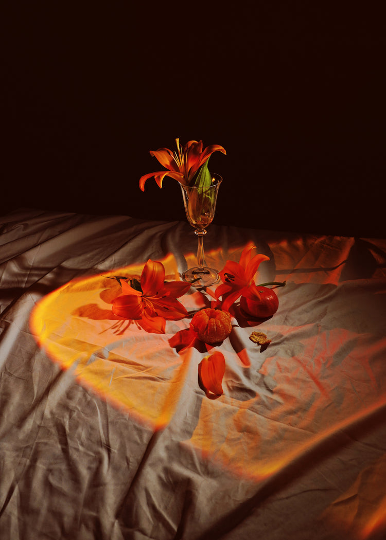 The Insomnia of the Flowers Poster showcases a still life composition with an orange lily in a glass on a table, scattered petals, two red apples, and warm lighting casting an orange glow and heart shape on the fabric surface.
