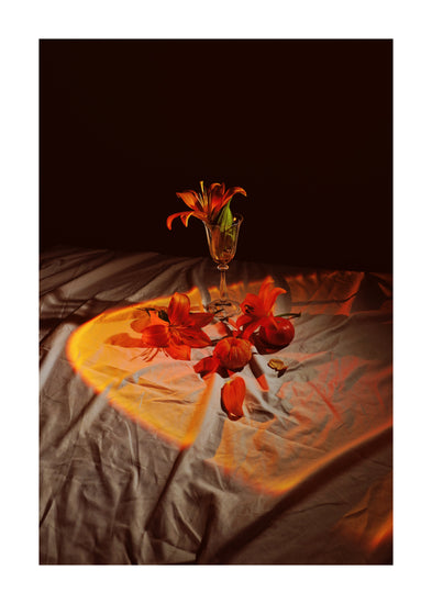 The Insomnia of the Flowers Poster depicts a still life with orange lilies—one in a glass vase and others scattered on fabric. Warm lighting creates an orange-yellow glow, casting dramatic shadows on a dark background to enhance vibrant colors and textures.
