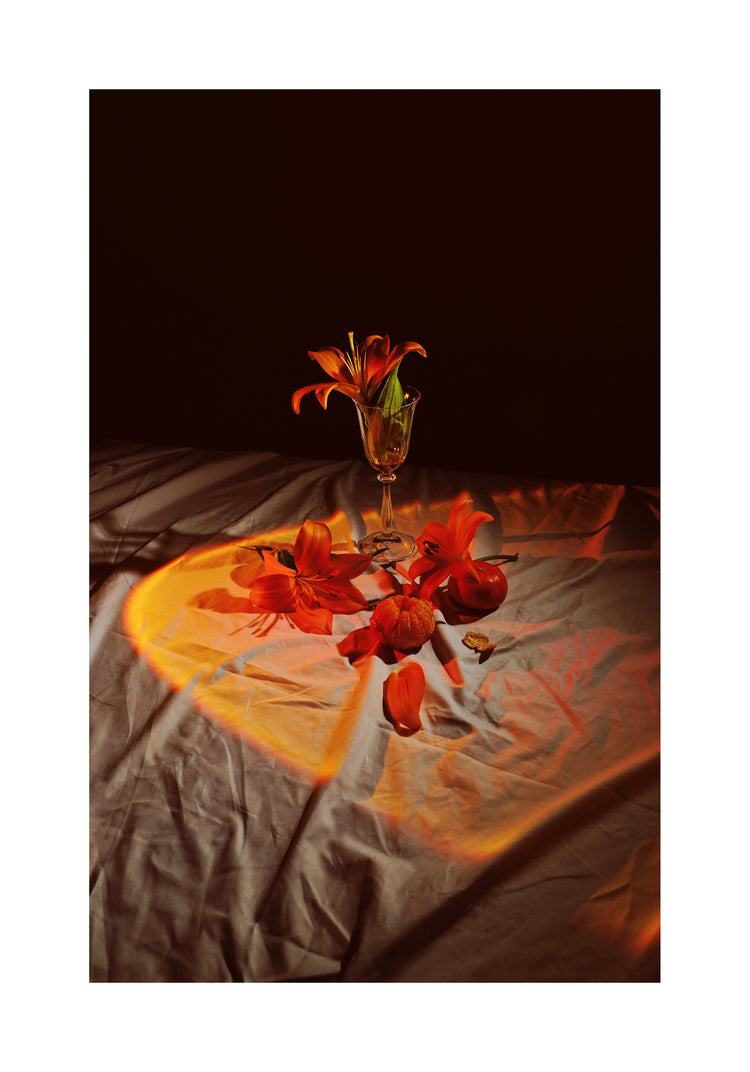 The Insomnia of the Flowers poster features a glass goblet filled with vibrant orange lilies, surrounded by scattered petals and mini pumpkins. Soft, warm light forms a heart shape on the cloth beneath, creating a cozy autumn mood against a dark backdrop.