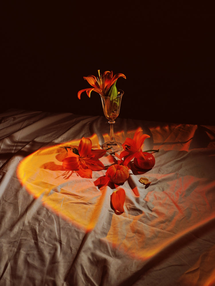 The Insomnia of the Flowers Poster showcases a still life with vibrant orange lilies in a tall glass on softly lit white cloth, surrounded by more lilies, an apple, and an orange, all enhanced by warm lighting that creates a cozy ambiance.