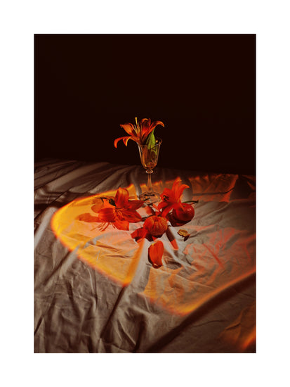 The Insomnia of the Flowers Poster showcases a still-life scene with orange lilies in a glass on draped cloth, scattered petals, and a small pumpkin. Soft, warm light creates dramatic shadows enhancing the compositions warmth and depth.