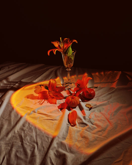 The Insomnia of the Flowers poster showcases a dimly lit still life with orange lilies in a tall glass, surrounded by small pumpkins and fallen petals on draped fabric, where warm light forms a heart-shaped glow on the cloth.