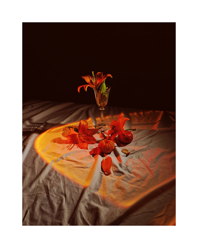 The Insomnia of the Flowers Poster depicts a glass vase with orange lilies on a dimly lit table, where scattered petals and two red apples are touched by a warm, circular light, crafting a dramatic and artistic scene.