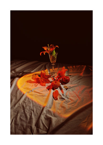 The Insomnia of the Flowers Poster showcases a vibrant still life with red-orange flowers in a glass vase against a dark backdrop, with petals and stems artfully scattered on softly lit, draped fabric for a warm, artistic effect.