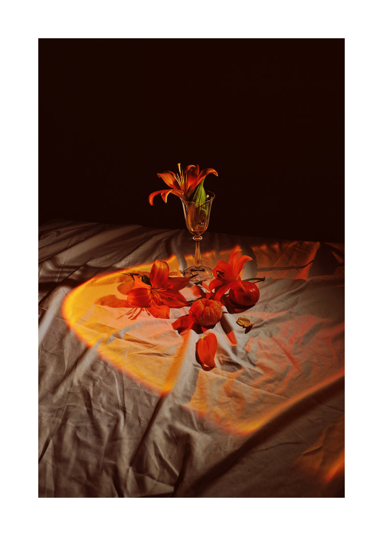 The Insomnia of the Flowers Poster showcases a vibrant still life with red-orange flowers in a glass vase against a dark backdrop, with petals and stems artfully scattered on softly lit, draped fabric for a warm, artistic effect.