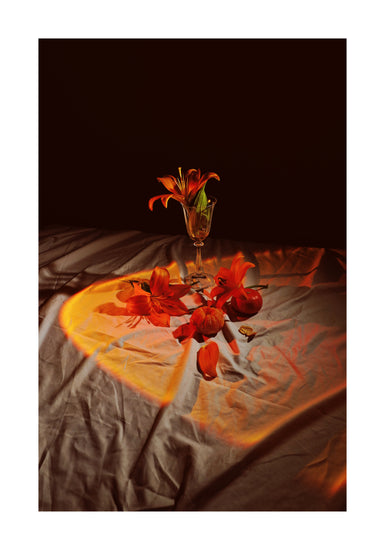 In the dim scene of the Insomnia of the Flowers Poster, vibrant orange lilies in a glass vase sit on a draped surface with scattered petals and stems; warm light casts a heart-shaped glow on the fabric.
