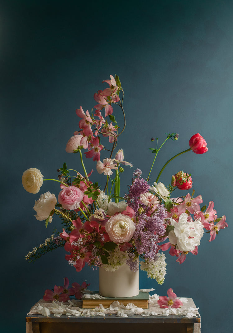 The Spring Ballet Poster showcases a vibrant flower arrangement in a white vase against a dark teal backdrop, featuring pink, red, and white blooms with green foliage and upward-reaching branches. The scene is set on a wooden surface with scattered petals.