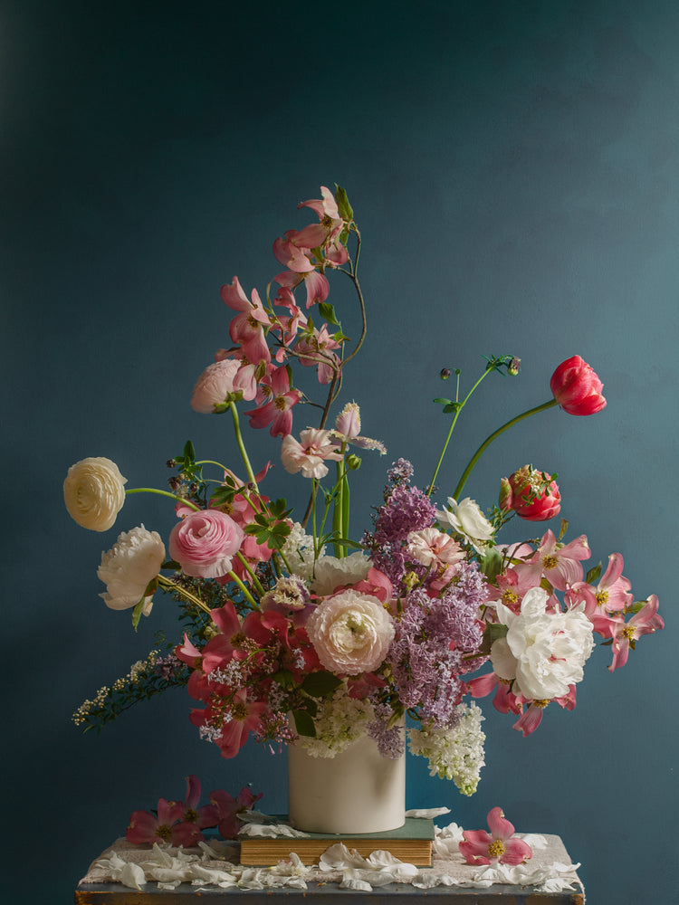The Spring Ballet Poster showcases a vibrant floral arrangement in a white vase, featuring pink, white, and purple peonies and ranunculus, all set against a moody dark teal background on a small table.