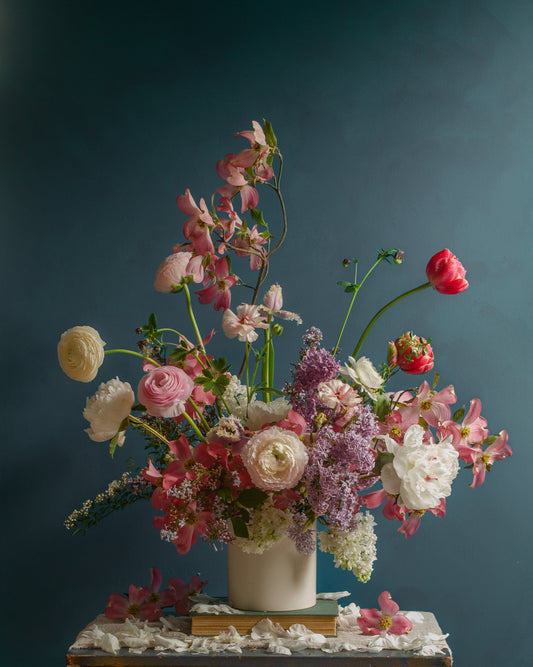 The Spring Ballet Poster showcases a vibrant floral arrangement in a white vase set against a dark teal background, highlighted by pink, white, and red roses and lilacs with scattered petals on the wooden surface below.