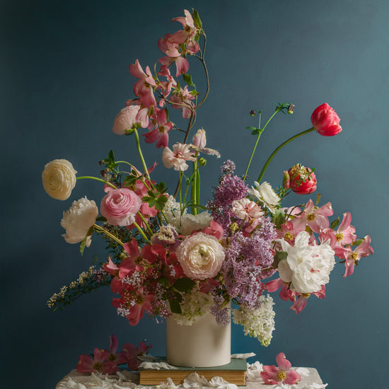 The Spring Ballet Poster displays a vibrant floral arrangement. It features pink peonies, white ranunculus, red poppies, and purple lilacs in a white vase on a lace-covered surface with scattered petals against a dark teal background.
